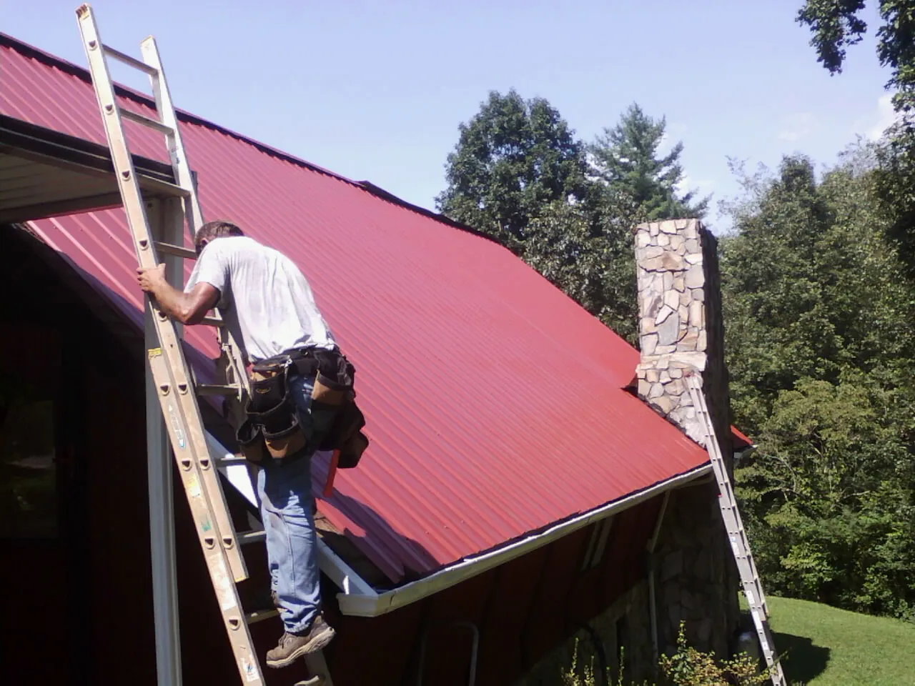 Roofer on a ladder beside a red metal roof and stone chimney — Soffit & Fascia Repair by Roofing Brothers in Elco